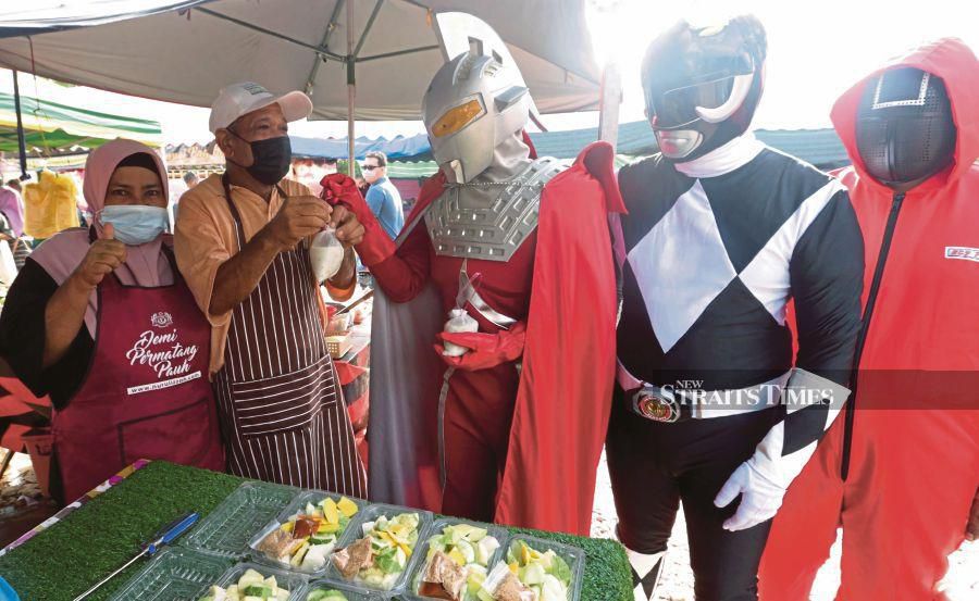 Mohamad Syukur Mohd Patthi (centre), in an Ultraman Seven costume, handing out ‘bubur lambuk’ to a trader at the Seberang Jaya Ramadan Bazaar in Butterworth, Penang, on Sunday. -NSTP/DANIAL SAAD