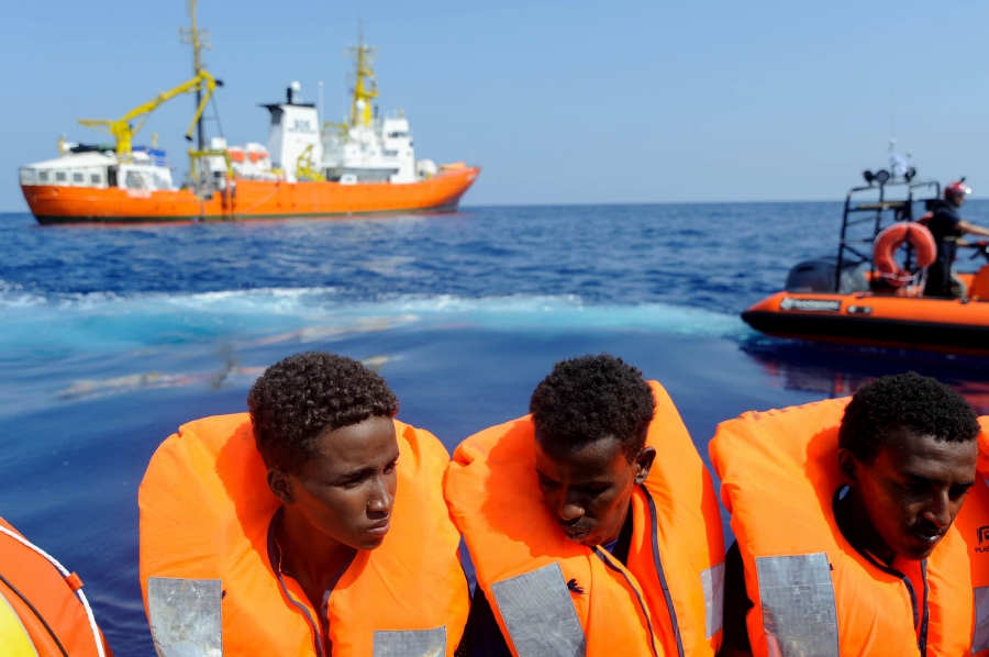 Migrants on a wooden boat are seen after receiving life jackets from crew members of the rescue boat Aquarius (background) on August 10, 2018 in the international waters off the Lybian coast. Photo by Guglielmo Mangiapane / SOS MEDITERRANEE / AFP 