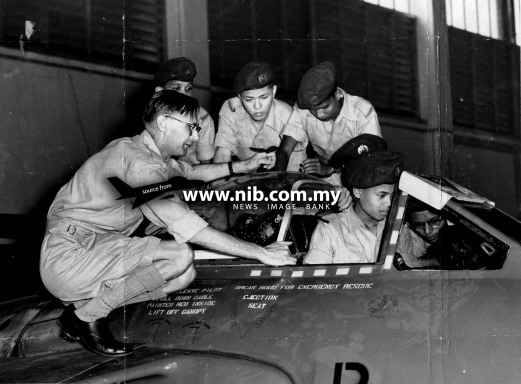 14 August 1956: Cadet Khairi Mohamed (in cockpit) of the M.A.T.C. finds it thrilling to make believe he is the pilot of this jet Venom fighter. On the left is F/Sgt. D. Goodinson, who is explaining the controls to him while his buddies look on