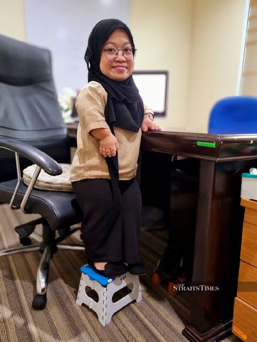 Emy Aridah Aripin standing at just 98-cm tall uused a small stool to reach the office table. - NSTP/Paul Mu